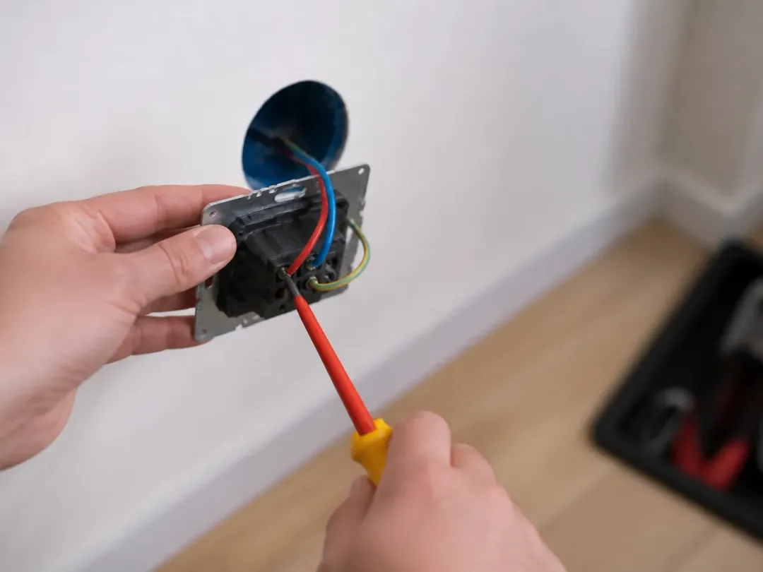 Mains tenant un mécanisme de prise sorti de sa boîte d’encastrement et serrant un fil avec un tournevis isolé