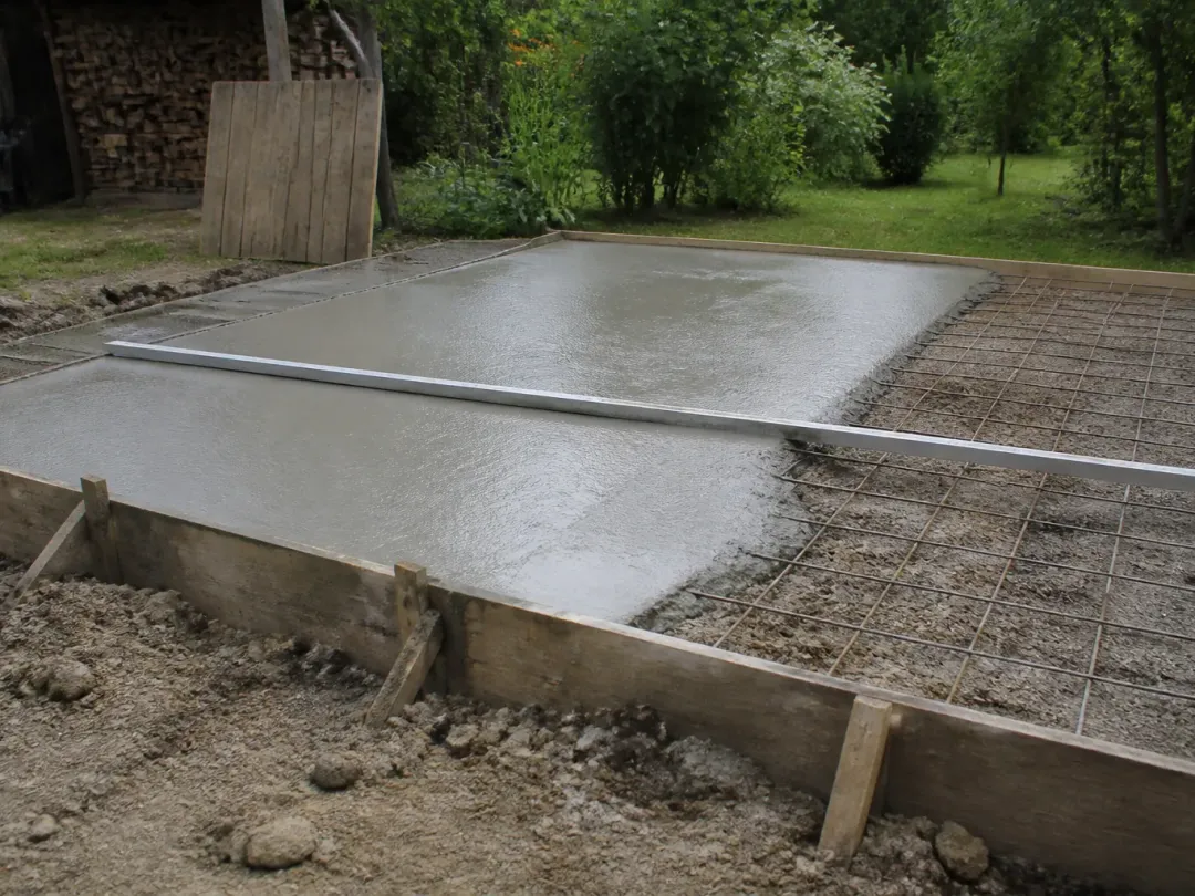 Dalle de béton fraîchement coulée dans un coffrage en planches, treillis soudé encore visible sur la partie non coulée