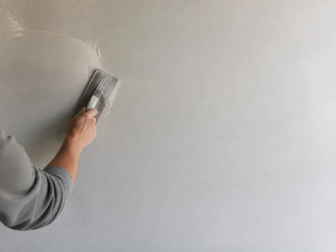 Main étalant une fine couche d’enduit blanc sur un mur avec une lisseuse inox