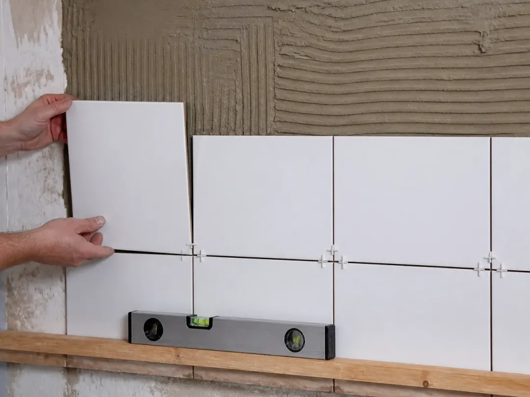 Mains posant des carreaux blancs sur un mur encollé au peigne, croisillons, tasseau et niveau à bulle en bas
