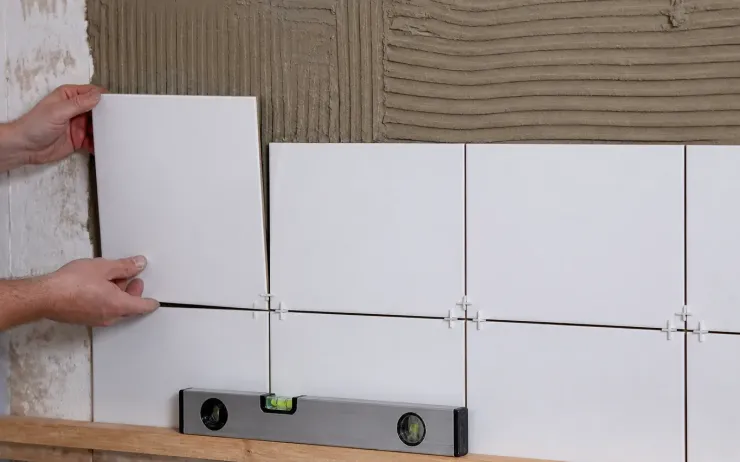 Mains posant des carreaux blancs sur un mur encollé au peigne, croisillons, tasseau et niveau à bulle en bas