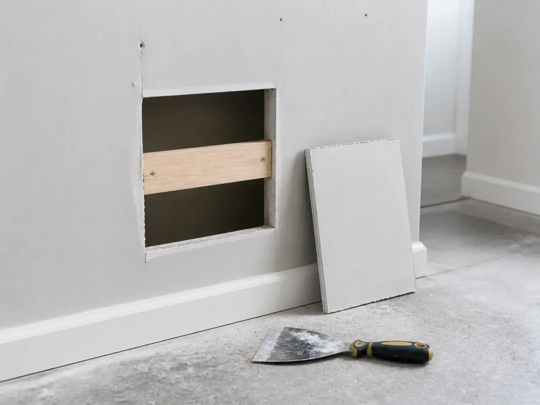 Découpe carrée dans une cloison en plaque de plâtre avec un tasseau de bois derrière, pièce de réparation et couteau posés au sol