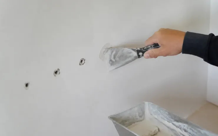 Main rebouchant des trous de cheville dans un mur blanc avec un couteau à enduire, auge d’enduit au premier plan