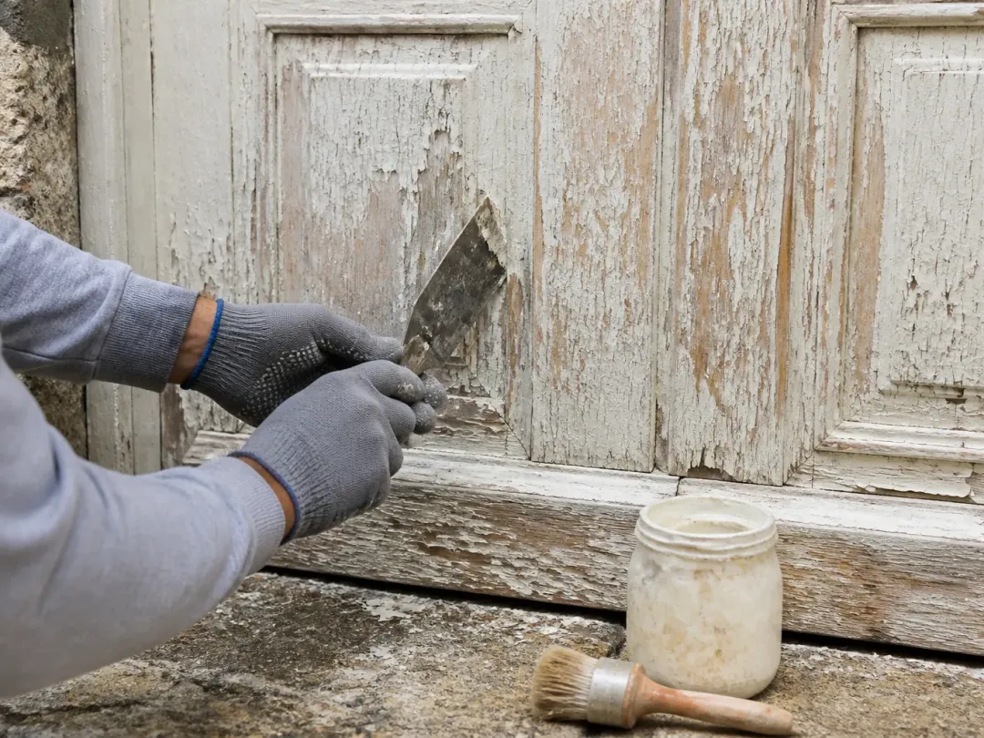 Mains gantées grattant au couteau une vieille peinture écaillée sur une porte en bois, pot de décapant au sol