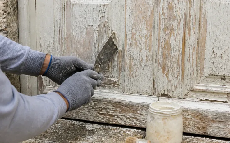 Mains gantées grattant au couteau une vieille peinture écaillée sur une porte en bois, pot de décapant au sol