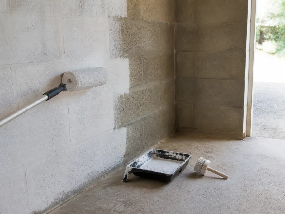 Mur en parpaings à moitié peint en blanc avec un rouleau à long manche, bac posé sur la dalle