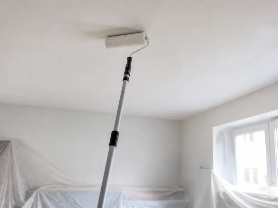 Rouleau sur manche télescopique appliqué contre un plafond blanc, meubles protégés par des bâches