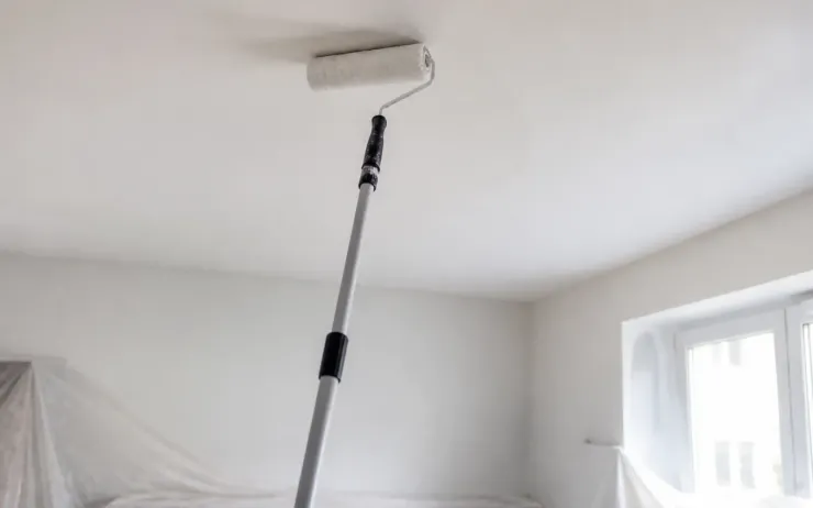 Rouleau sur manche télescopique appliqué contre un plafond blanc, meubles protégés par des bâches