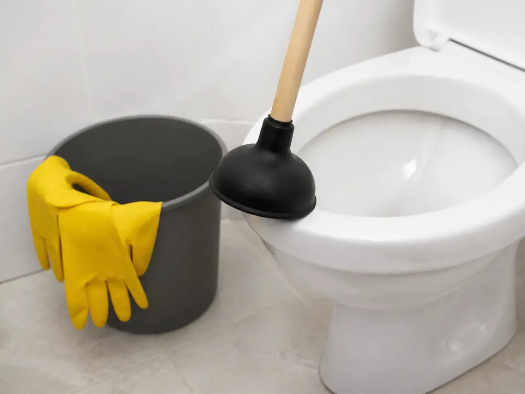 Ventouse à manche en bois devant une cuvette de WC, seau et gants de ménage jaunes