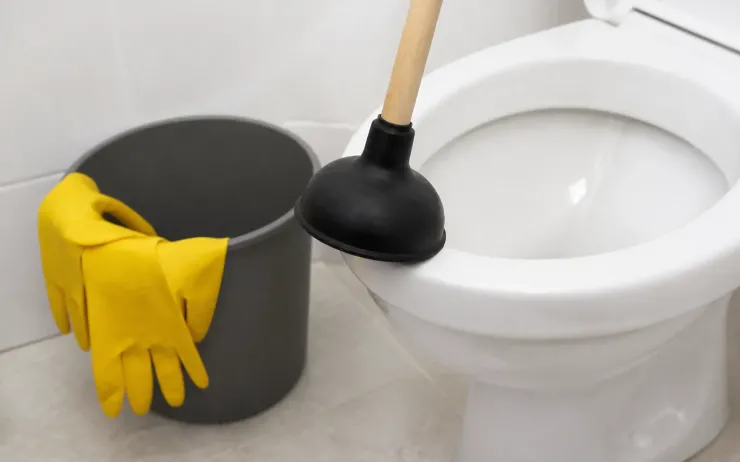 Ventouse à manche en bois devant une cuvette de WC, seau et gants de ménage jaunes