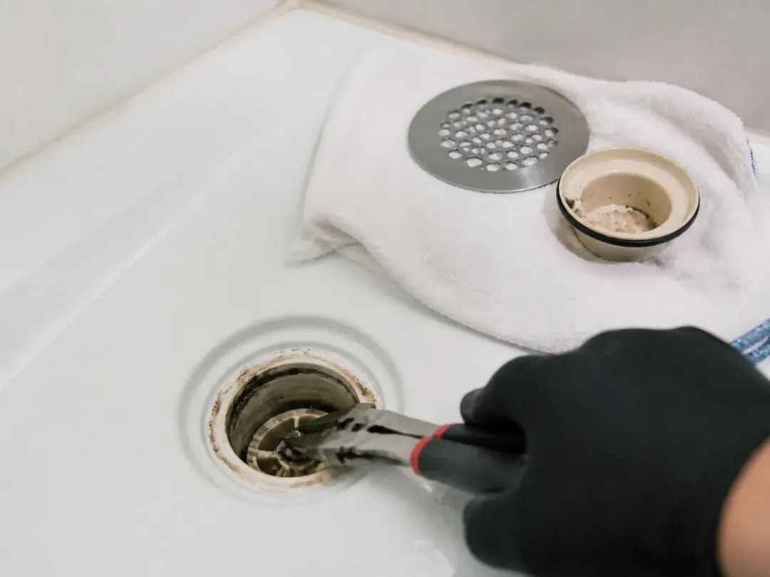Main gantée retirant les cheveux d'une bonde de receveur de douche à la pince, grille posée sur une serviette