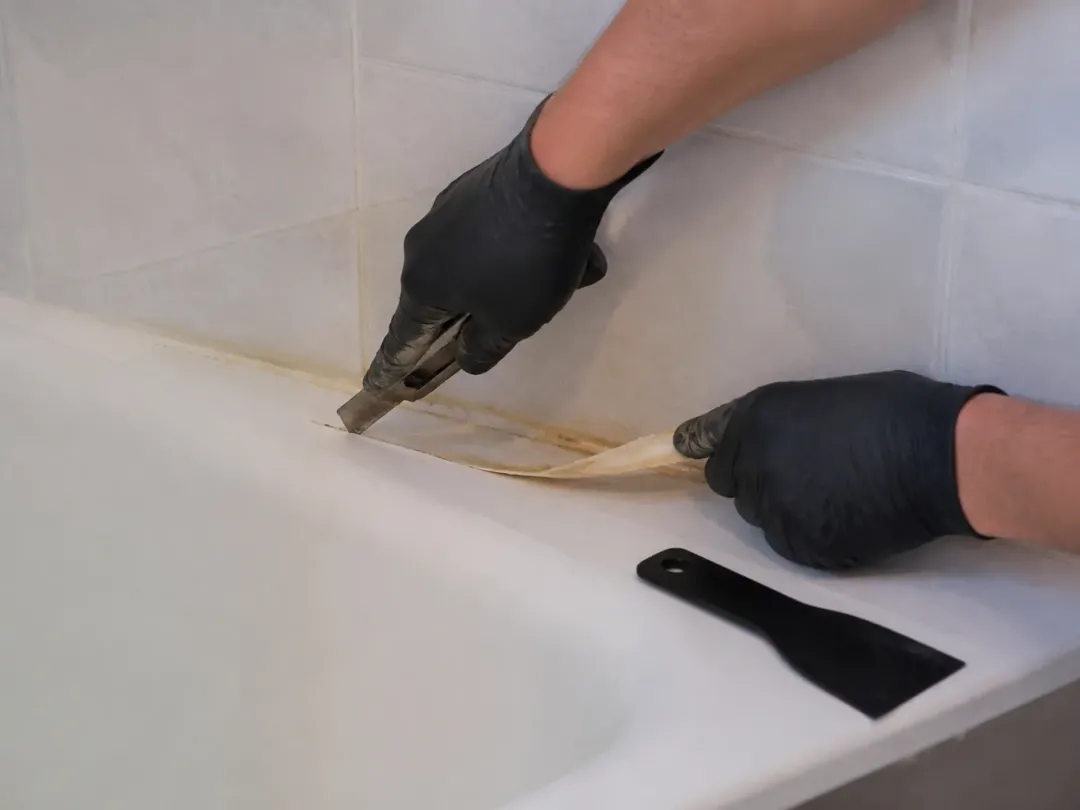 Mains gantées retirant au cutter un vieux joint silicone jauni au bord d’une baignoire, grattoir en plastique à côté