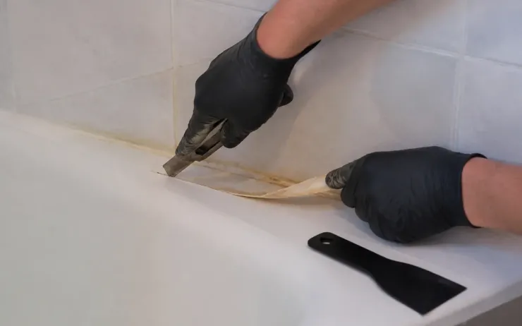 Mains gantées retirant au cutter un vieux joint silicone jauni au bord d’une baignoire, grattoir en plastique à côté