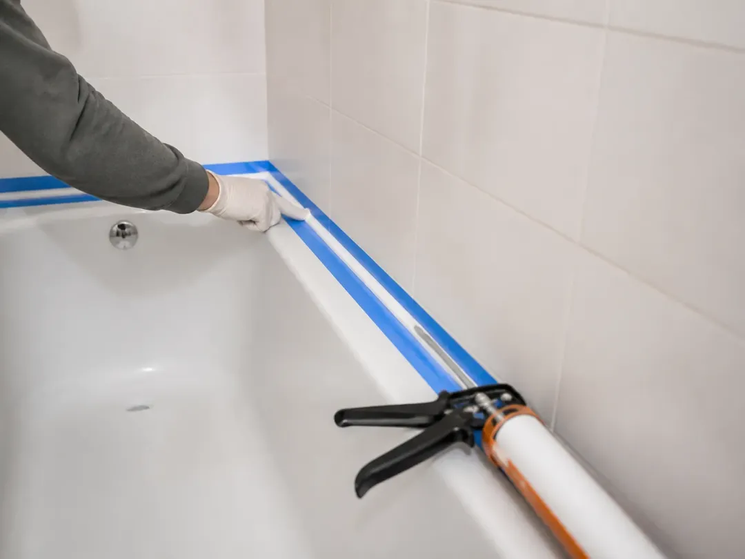 Joint silicone en cours de lissage au bord d’une baignoire, ruban de masquage bleu et pistolet à cartouche posés dessus