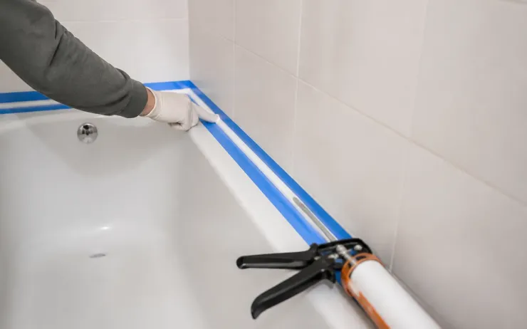 Joint silicone en cours de lissage au bord d’une baignoire, ruban de masquage bleu et pistolet à cartouche posés dessus