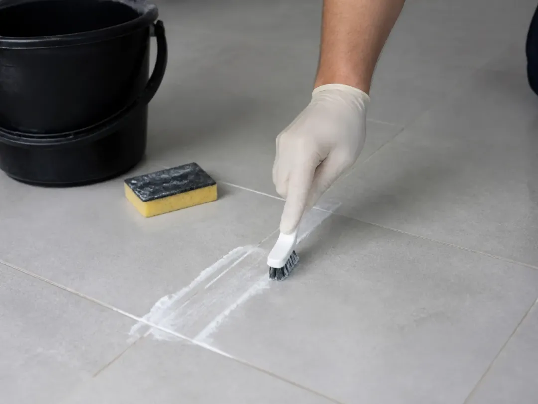 Main gantée brossant un joint de carrelage gris clair enduit de pâte blanche, éponge et seau à côté
