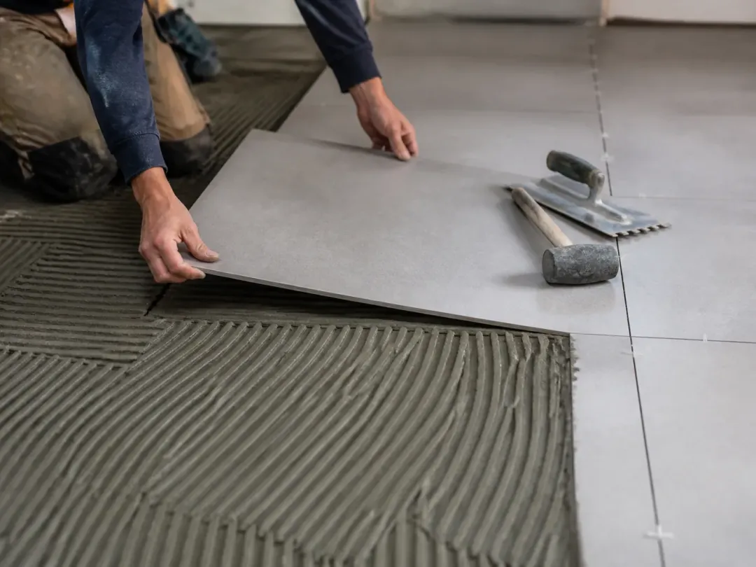 Personne à genoux posant un grand carreau gris sur un mortier-colle peigné, spatule crantée et maillet à côté