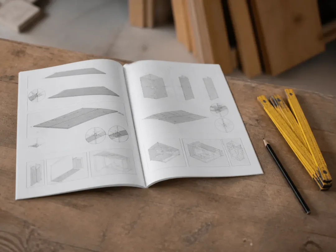 Notice technique à schémas ouverte sur un établi, à côté d’un mètre pliant jaune et d’un crayon